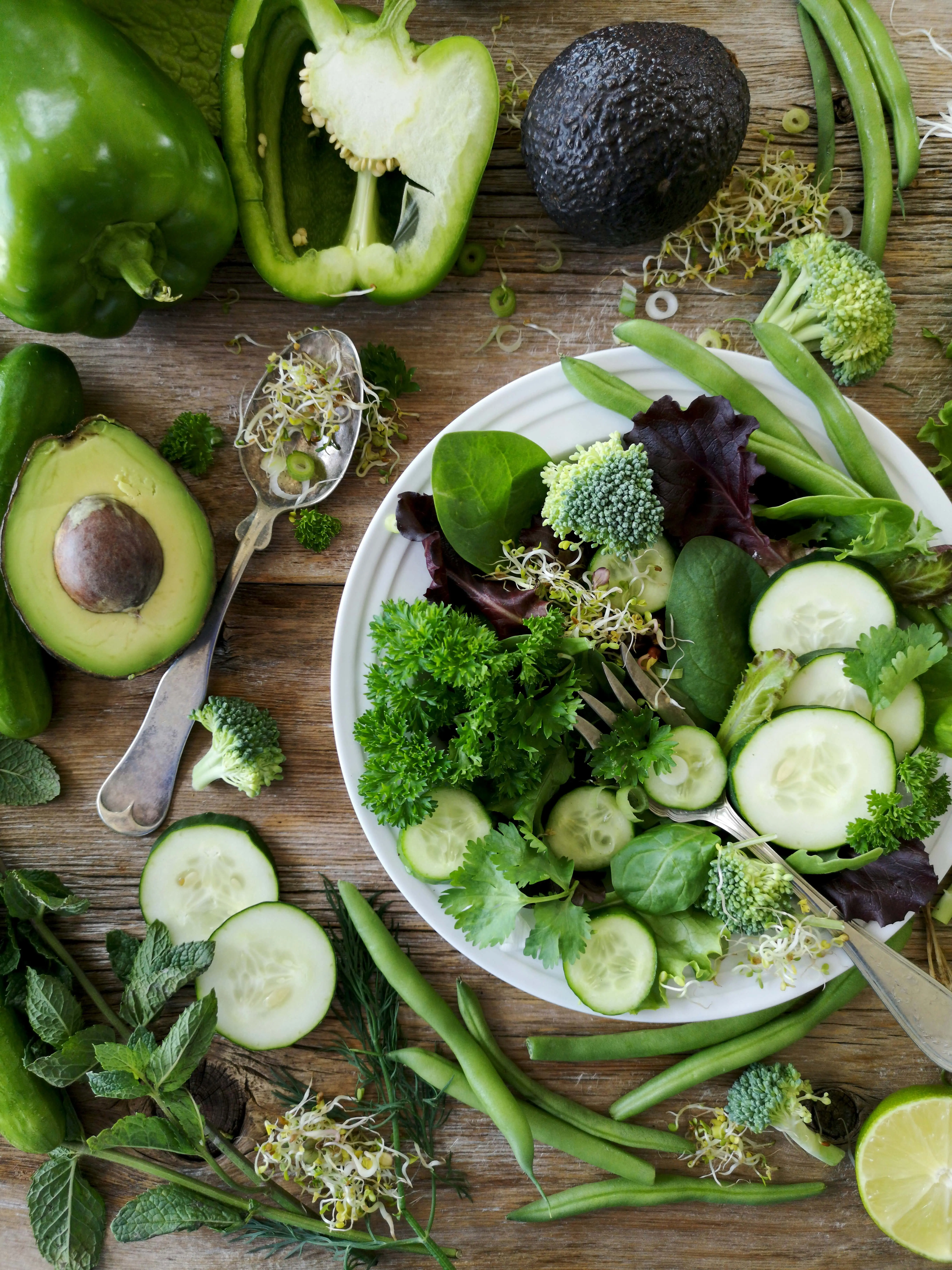 La imagen muestra un tazón blanco lleno de una ensalada de verduras verdes. Las verduras parecen ser principalmente lechuga romana, espinacas y rúcula. También hay algunas rodajas de tomate y pepino. El tazón está colocado sobre una mesa de madera.